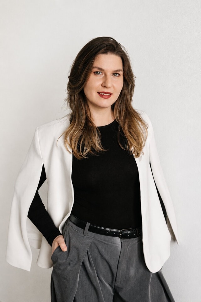 Ksenia Gavrilova standing in a white cape jacket against a pale studio background.
